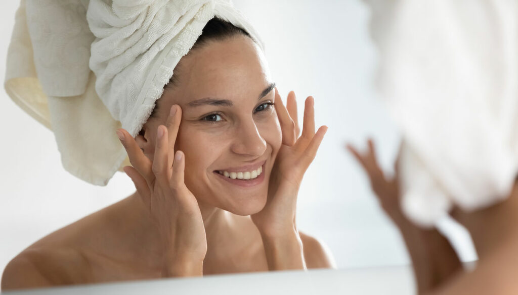 woman with her hair in a towel looking at her face in the mirror, chemical peel