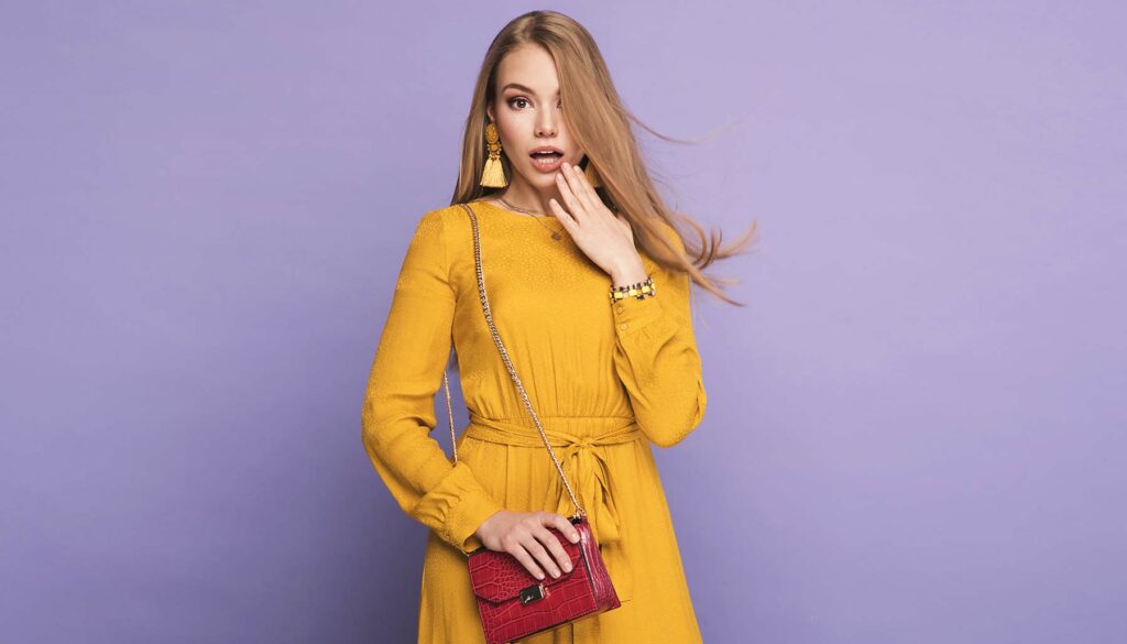 woman against a purple background wearing a fashionable yellow dress and holding a red purse