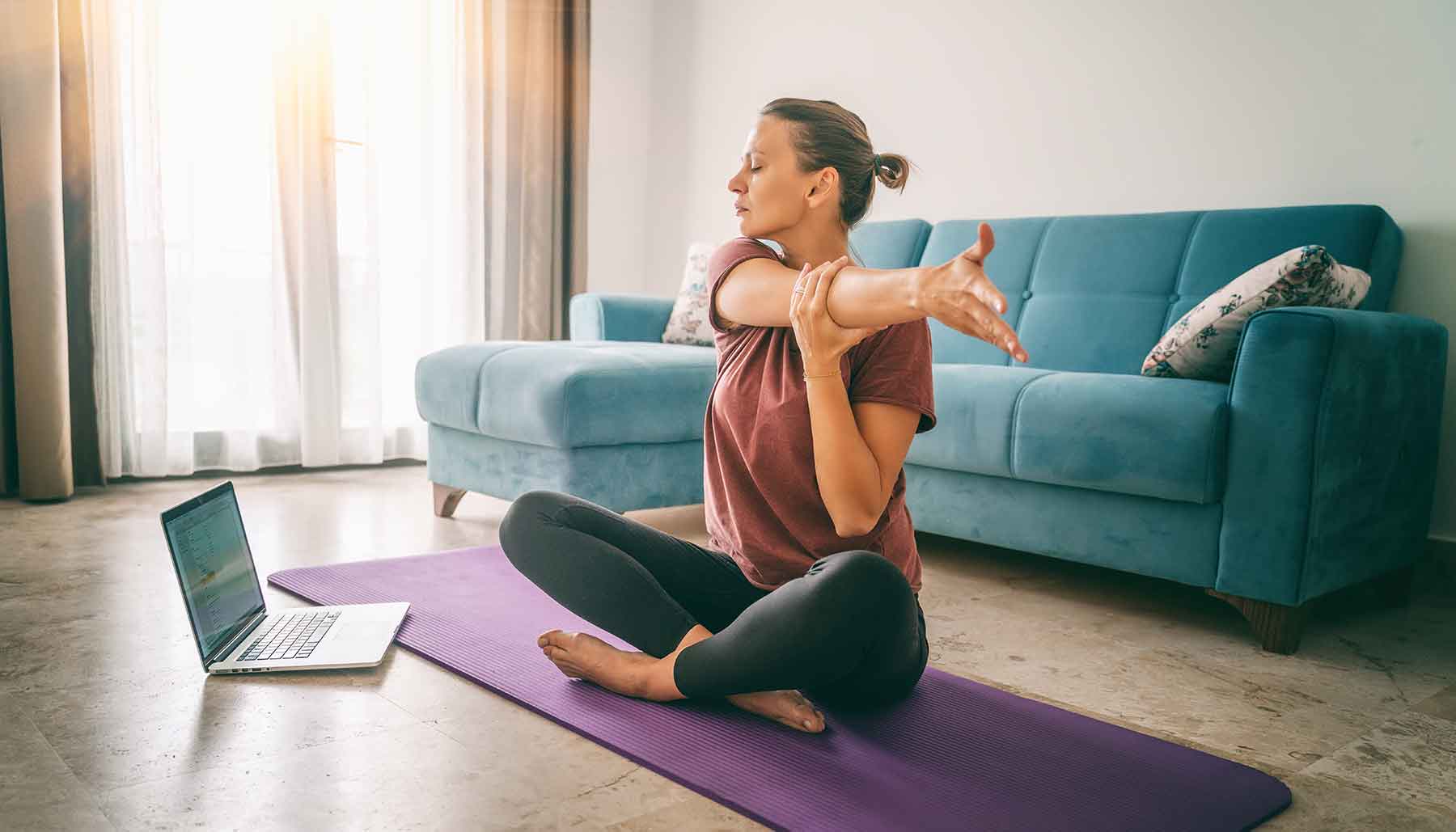 woman sitting on yoga mat in living room with laptop, stretching