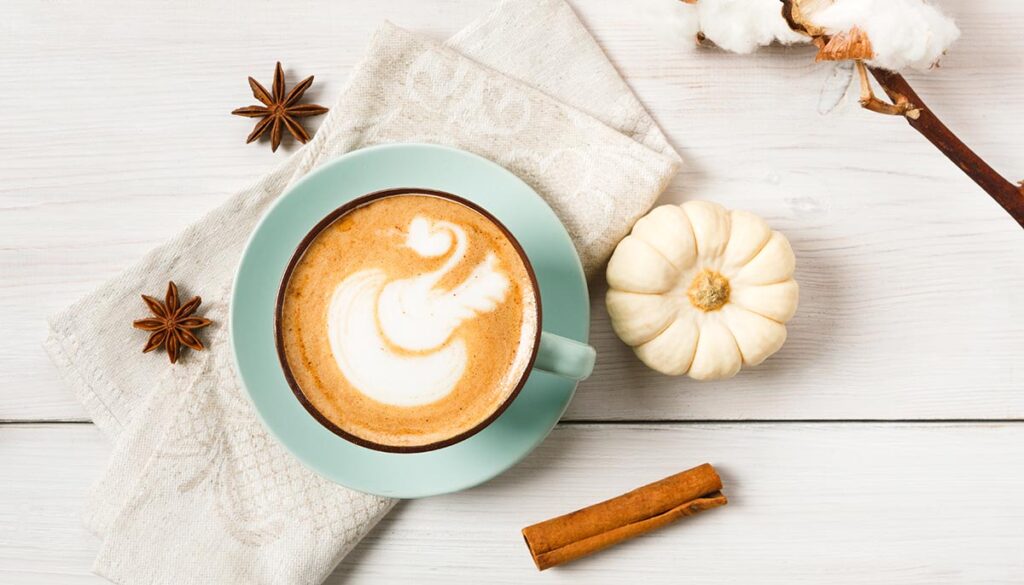 Pumpkin spice latte. Blue coffee cup with creamy foam, cinnamon sticks, cloves and small yellow pumpkins at white wood background. Autumn fall hot drinks, cafe and bar concept, top view