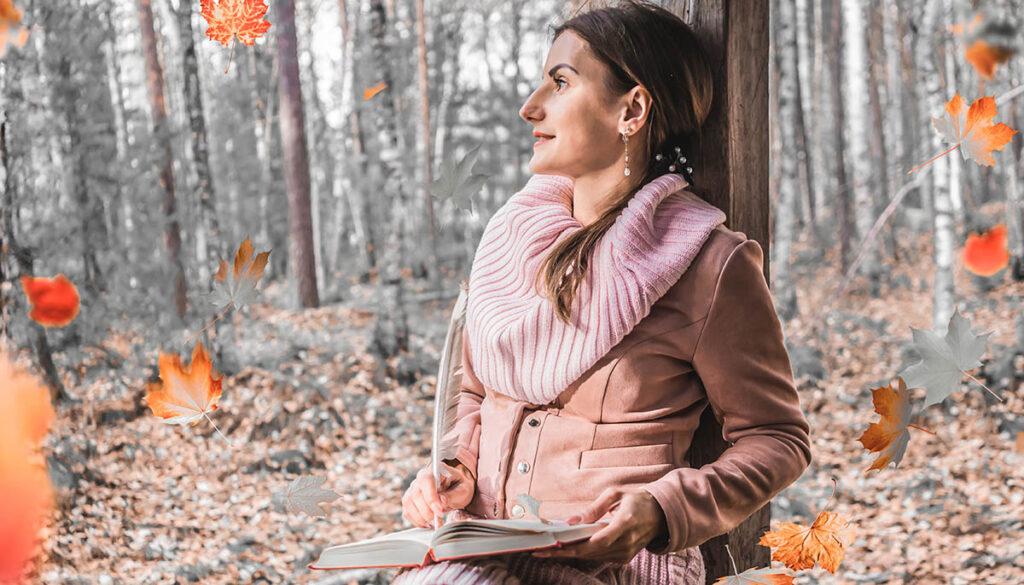 A woman holding a journal and pencil looks out on the autumn woods