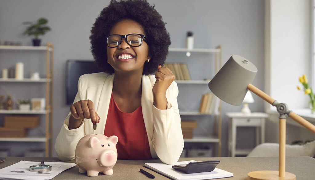 Money saving for shopping concept. Lovely satisfied, excited happy young african american woman in eyewear with yes gesture putting coin into piggybank because of income exceeds expenses significantly