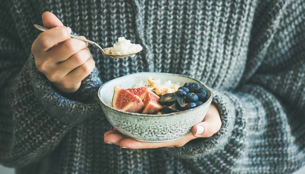 woman in cozy gray sweater eating healthy breakfast of rice and coconut with blueberries and guava