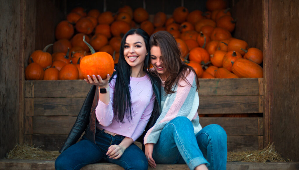 two women smiling and sitting in front of many pumpkins fall