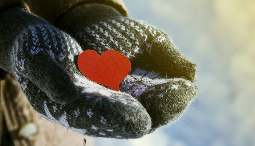 Hands wearing mittens, holding out a paper heart. Representing the concept of holiday generosity.