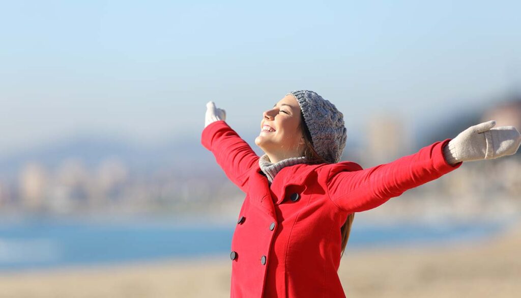 Happy woman wearing a red jacket breathing fresh air and raising arms on the beach in a sunny day of winter