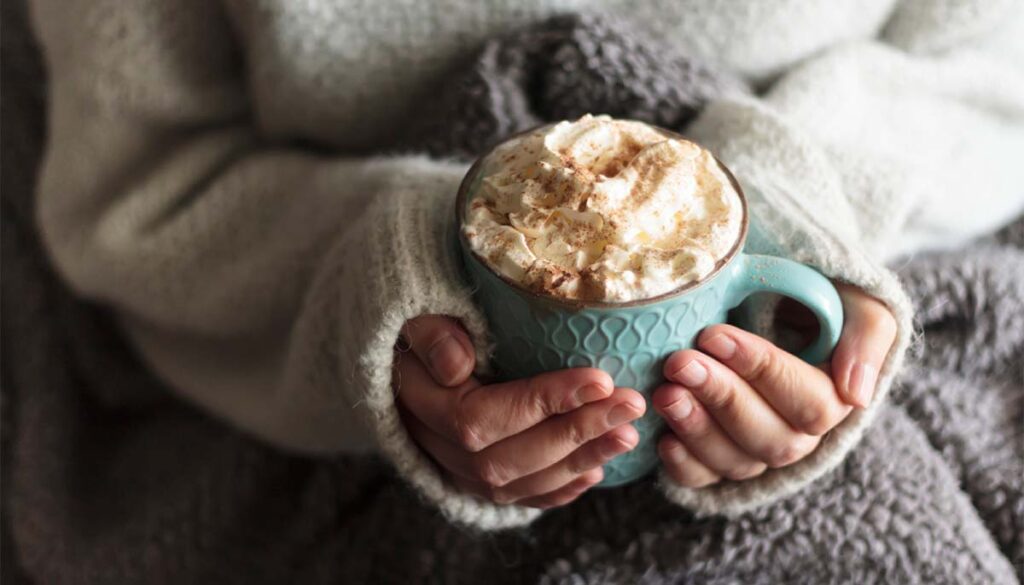 Woman with blanket warming her hands in mug of hot drink with whipped cream
