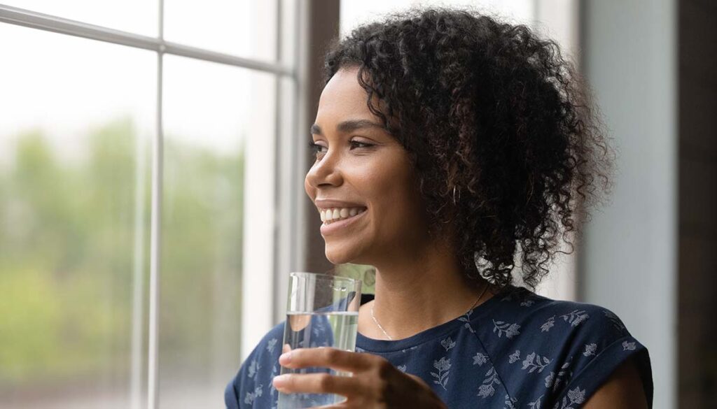 Smiling millennial woman looking in distance and holding glass of water 