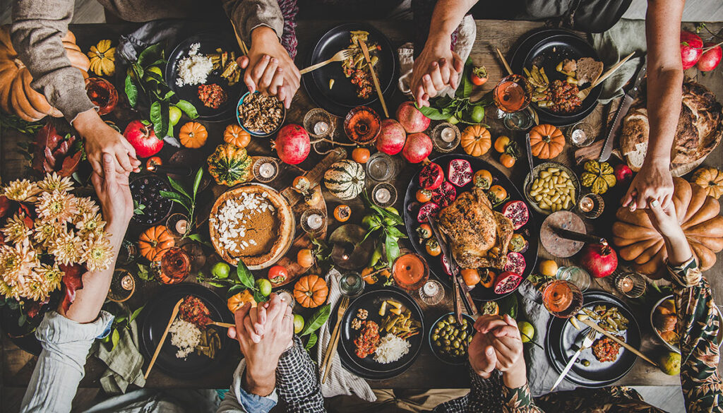 Family praying holding hands at Thanksgiving table. Flat-lay of feasting peoples hands over Friendsgiving table with Autumn food, candles, roasted turkey and pumpkin pie over wooden table, top view