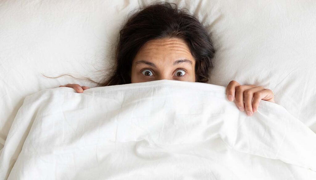 woman with eyes wide open with surprise hiding lower face behind blanket in bed
