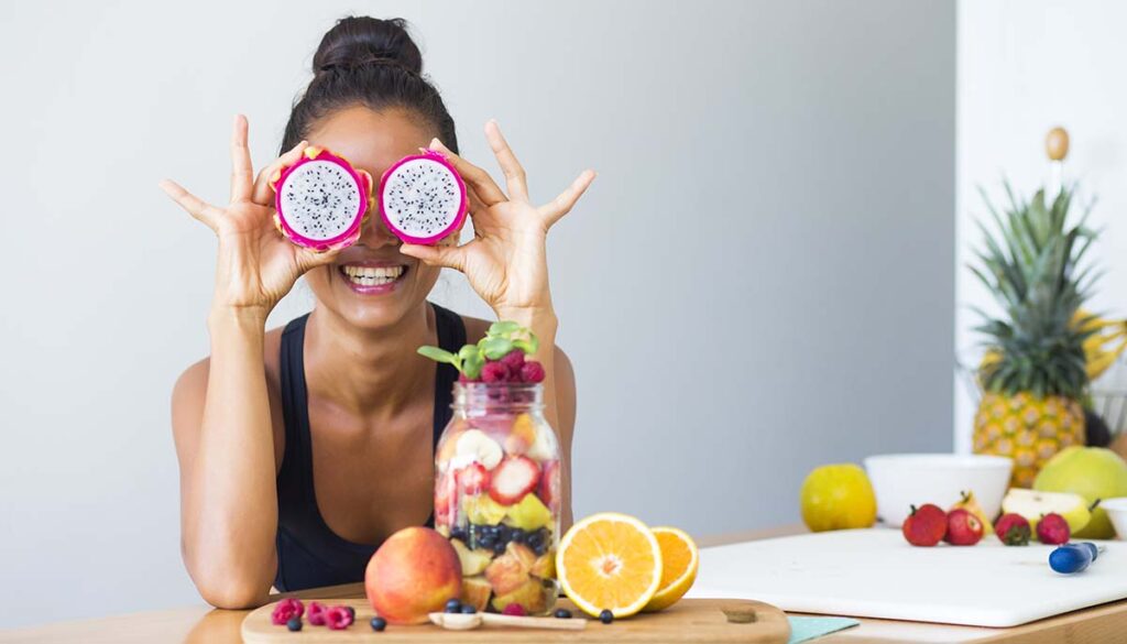 woman smiling and holding two halves of dragon fruit in front of her eyes with various fruits on the table