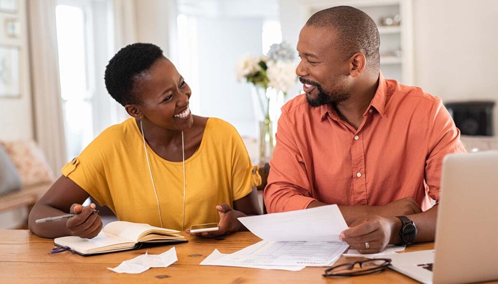 Cheerful mature couple sitting and managing expenses at home. Happy african man and woman paying bills together and managing budget. Black smiling couple checking accountancy and bills while looking at each other.