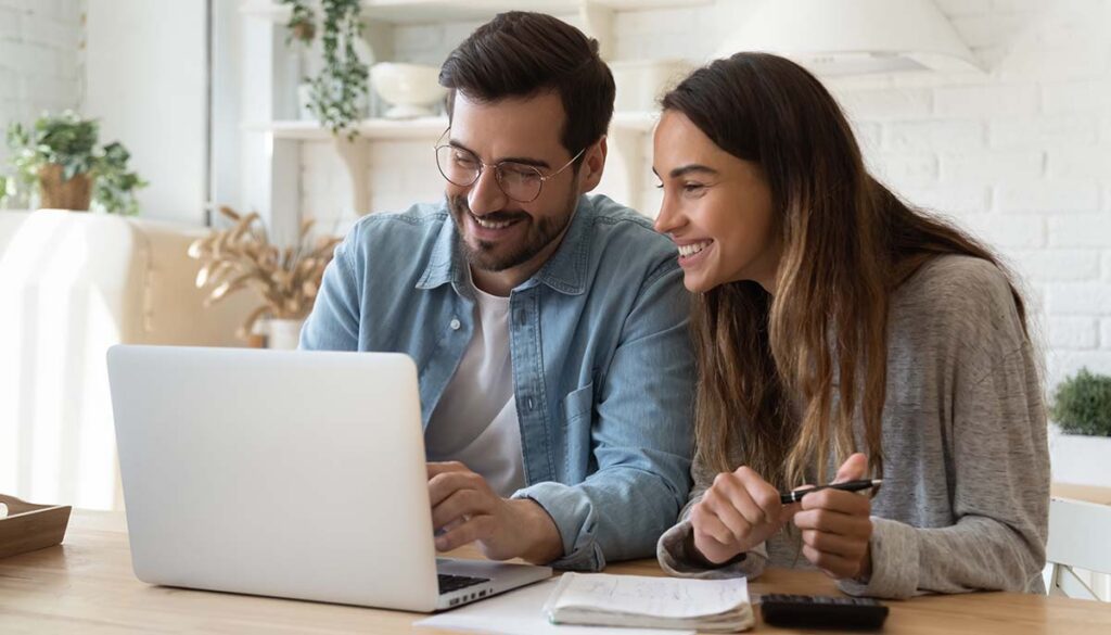 Happy young couple husband and wife using laptop computer looking at screen pay bills online in app calculate mortgage investment payment on website planning budget discuss finances sit at home table