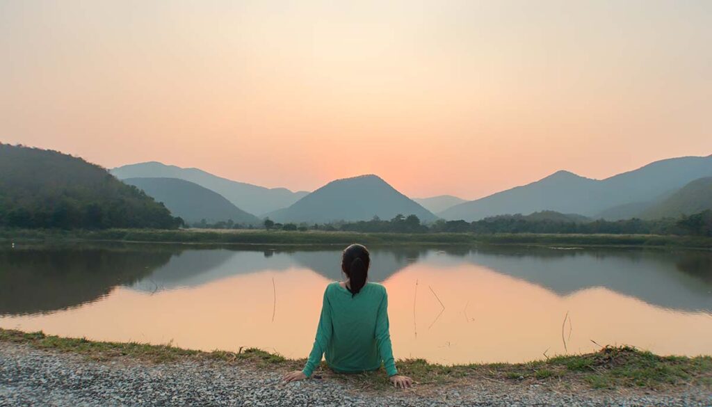 back of lone woman looking out into a lake with mountains reflecting on water at sunset