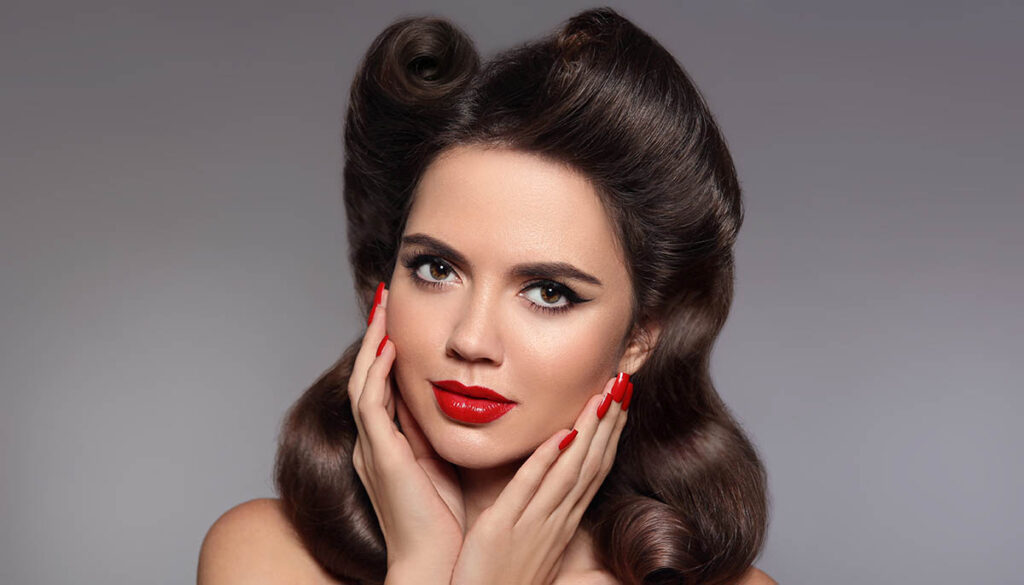 Pin up hairstyle. Beautiful 50s girl holding her cheeks with red lips makeup and manicured nails looking at camera. Expressive facial expressions. Beauty fashion studio photo.