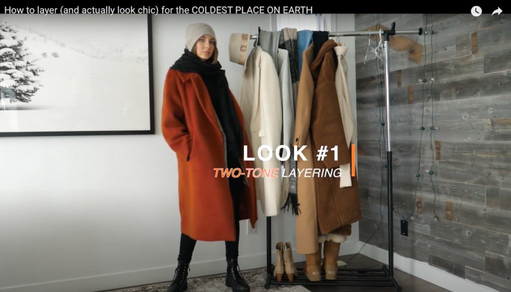 A screenshot from Amber Scott's Youtube video on layering, she's wearing a long orange coat with a black scarf and tan beanie. 