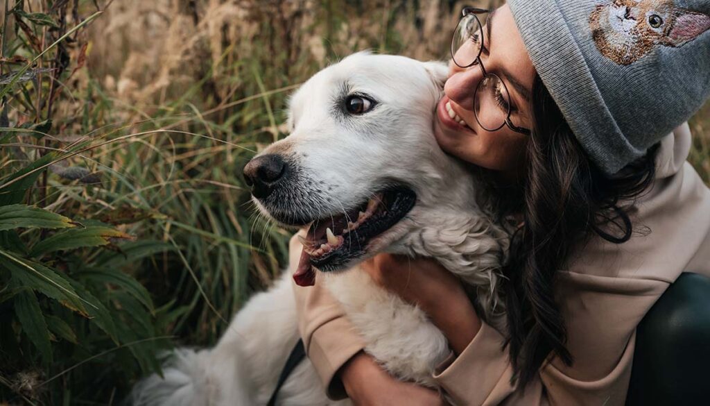 brunette in glasses and beanie hugging golden retriever in nature