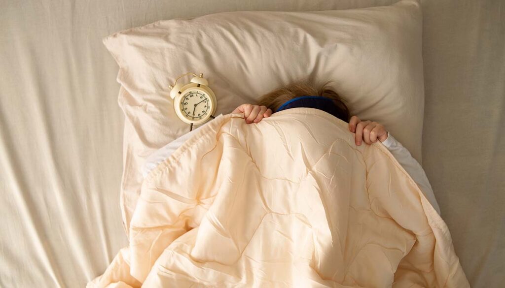woman in bed covering her face with the blanket with her alarm clock on her pillow