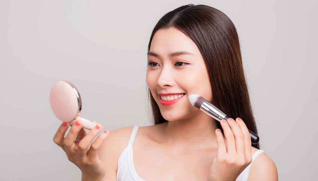 smiling woman looking into compact mirror and putting on blush