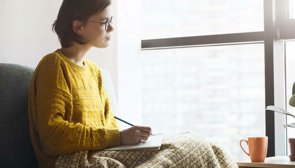 woman writing in journal at window, drinking coffee or tea in mug