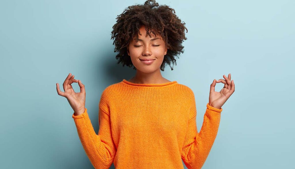 young woman in orange sweater meditating with eyes closed against light blue backdrop 
