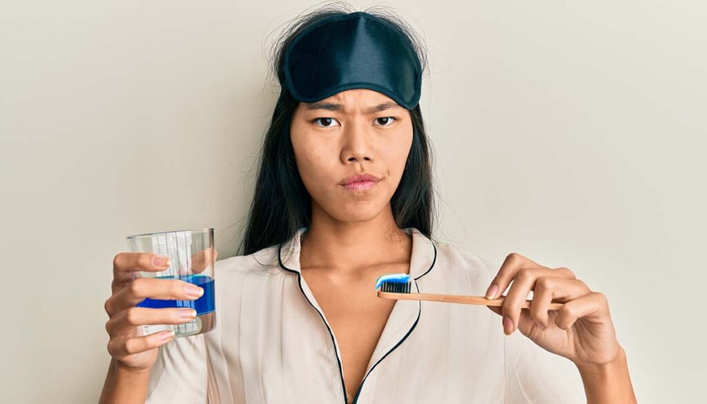 young woman in pjs with sleep mask on head brushing her teeth, gargling mouthwash, and looking irritated 