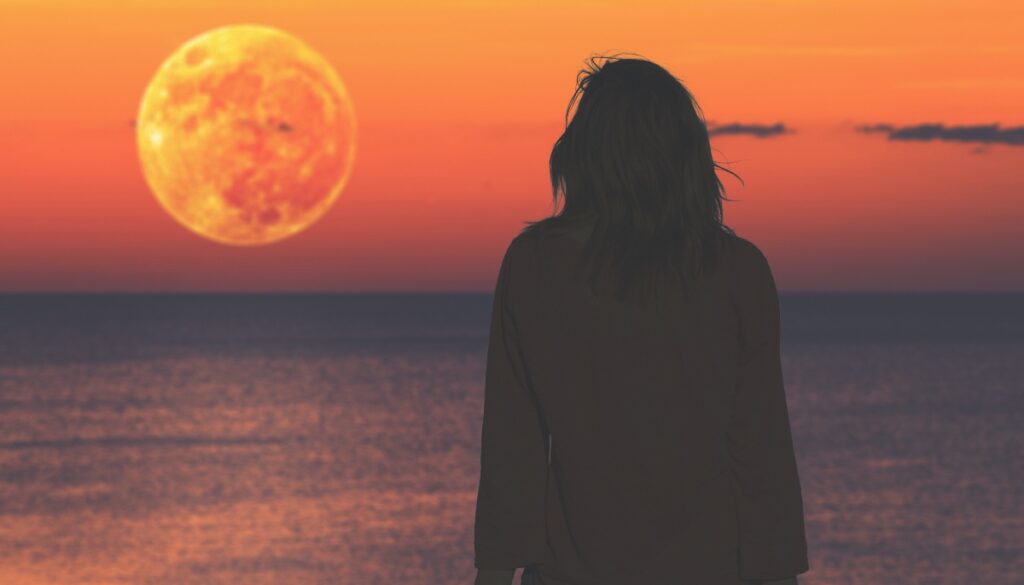 shadowy figure looks out at an orange moon over the ocean