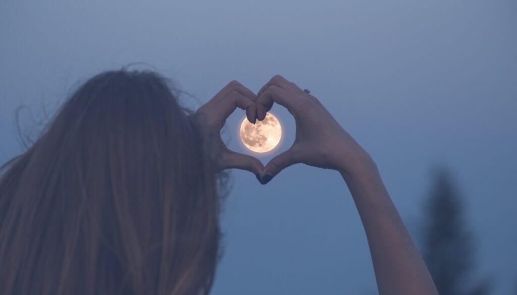 person forms a hand heart around the full moon