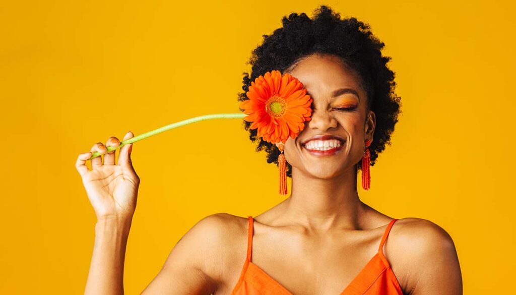 happy woman holding orange gerber daisy to face, wearing an orange dress, on an orange background