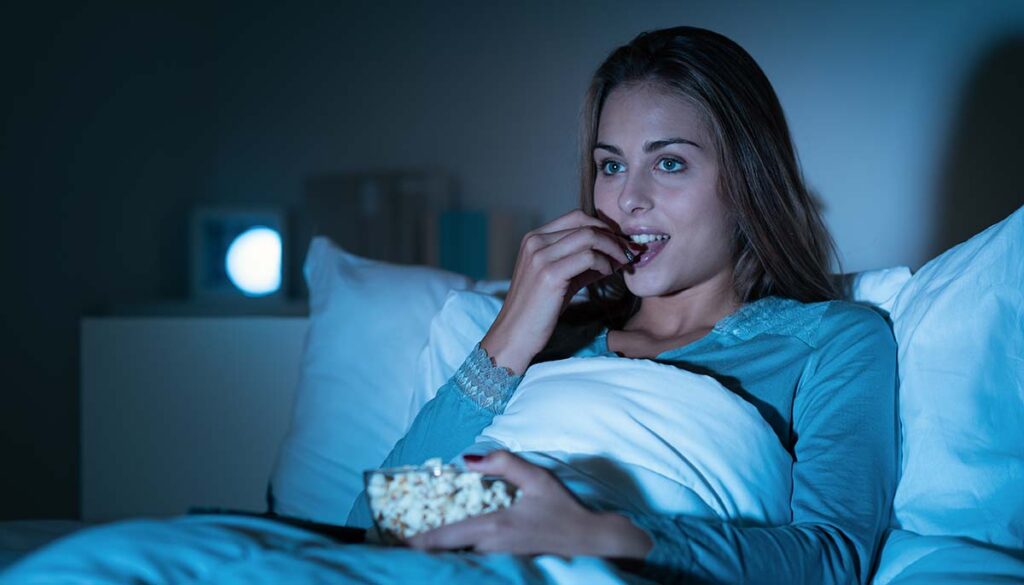 woman watching tv in bed while eating popcorn