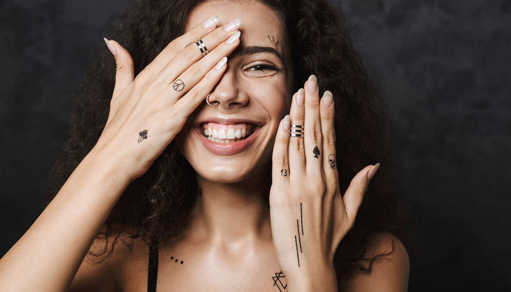 Image of young cheerful woman with tattoo smiling and covering her face isolated over black background