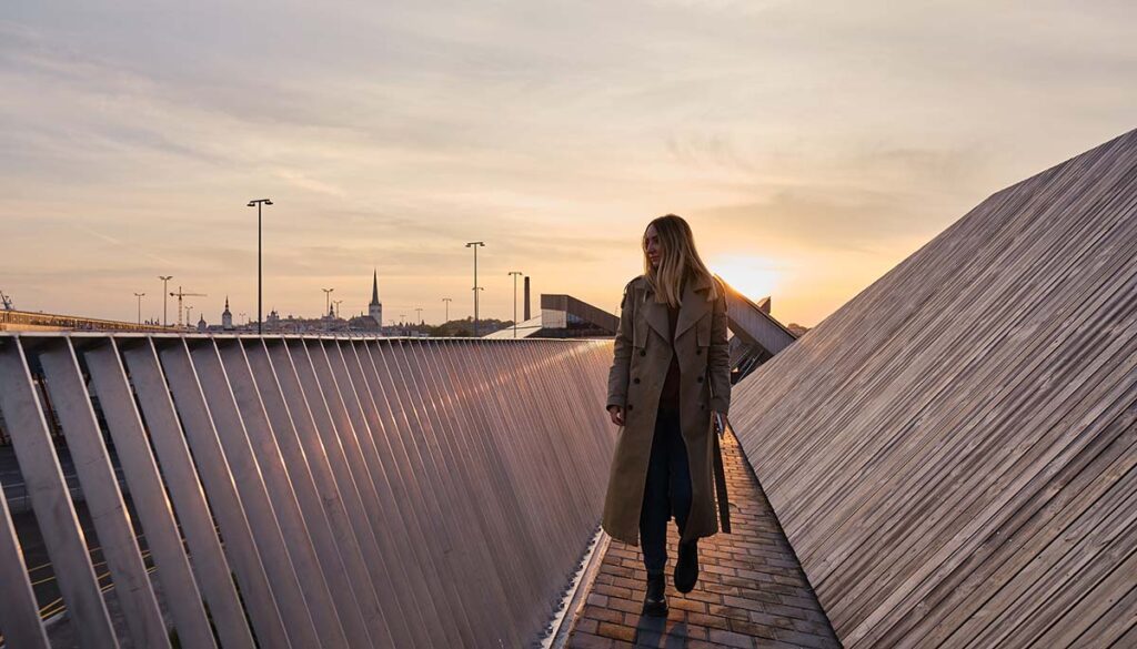 A young woman in a trench coat stands on a modern bridge in the city and looks at the sunset.