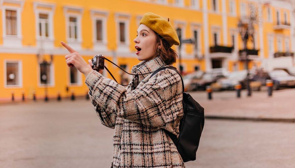 enthusiastic, female tourist taking photos of a city while in tweed jacket and yellow beret with yellow building behind her