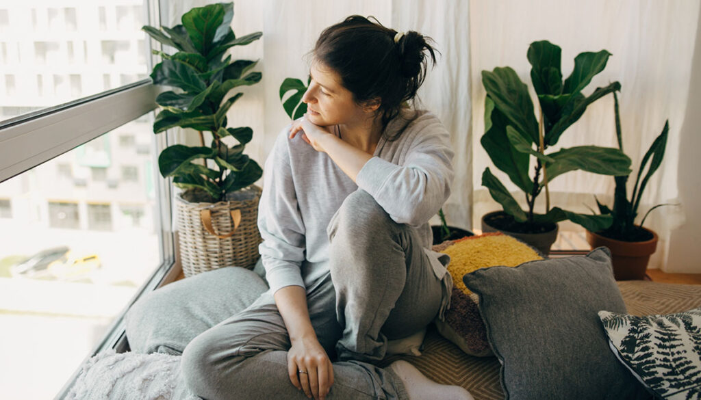 Sad hipster girl sitting at home during quarantine and looking at window. Stay home stay safe. Isolation at home to prevent virus epidemic. Young woman relaxing in modern room