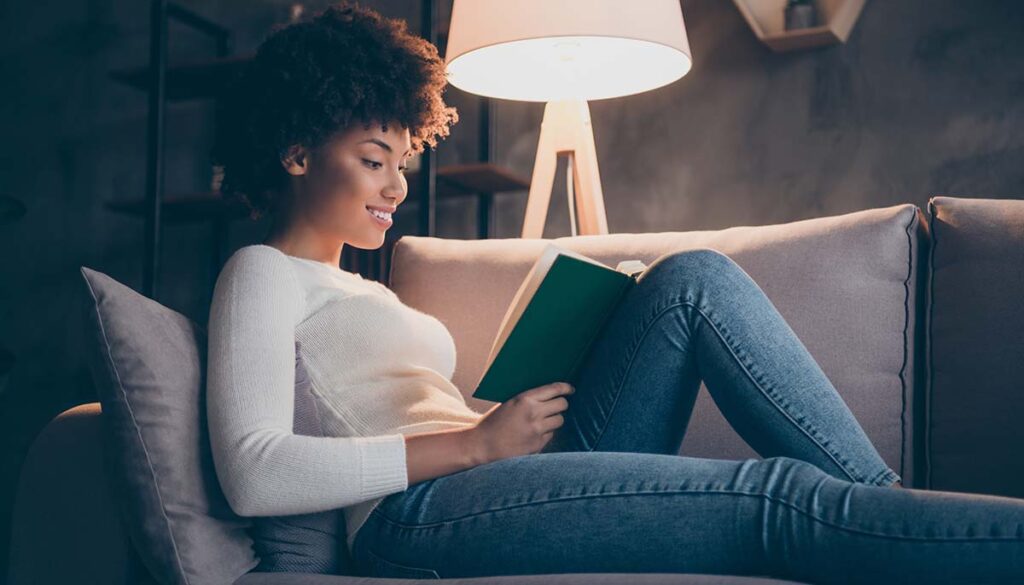 woman holding favorite historic novel reading excited lying cozy couch wear casual sweater jeans outfit evening living room indoors