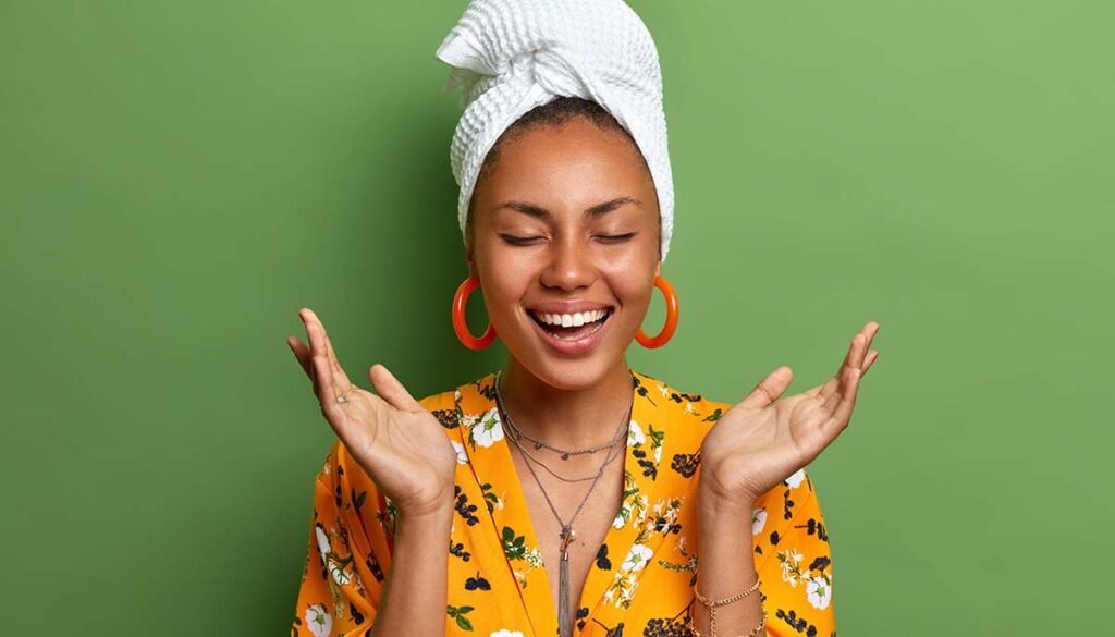 woman spreads palms laughs happily closes eyes wears earrings bath towel wrapped on head with glowing skin