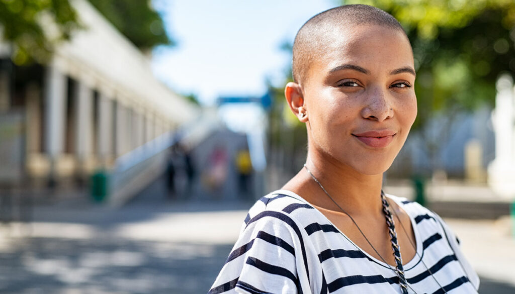 Portrait of young happy bald woman on city street looking at camera. Confident stylish girl outdoor with copy space. Proud and satisfied black curvy woman standing on street.