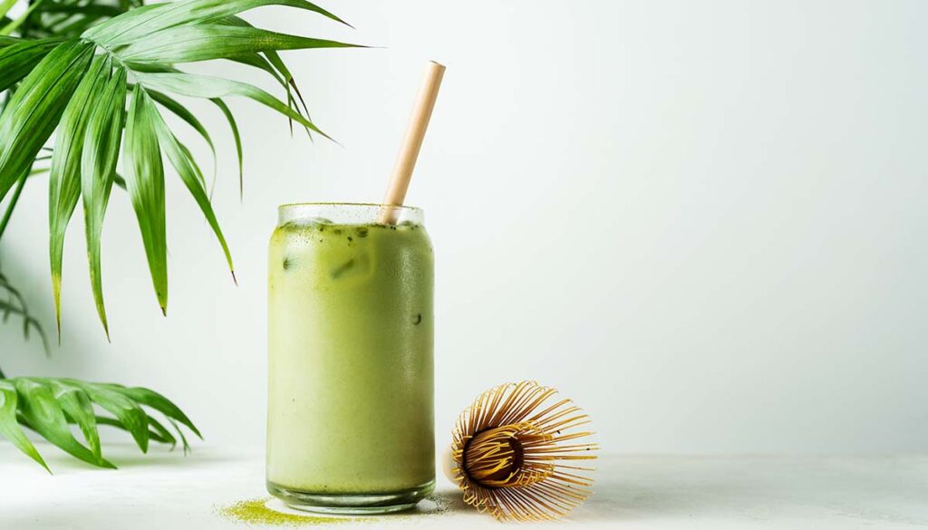 Making Japanese iced matcha latte, green tea with milk, soy milk, traditional matcha tools, with bamboo straw in glass on white background.