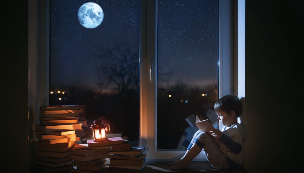 Little boy reading by a large window under the light of the moon