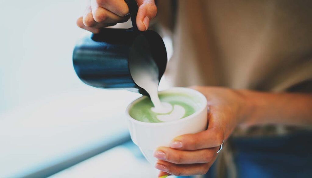 Closeup photo of a woman's hands pouring a milk into a freshly made matcha tea. Preparing japanese green tea with a milk. Female barista making matcha latte. Matcha green tea with a hot milk.
