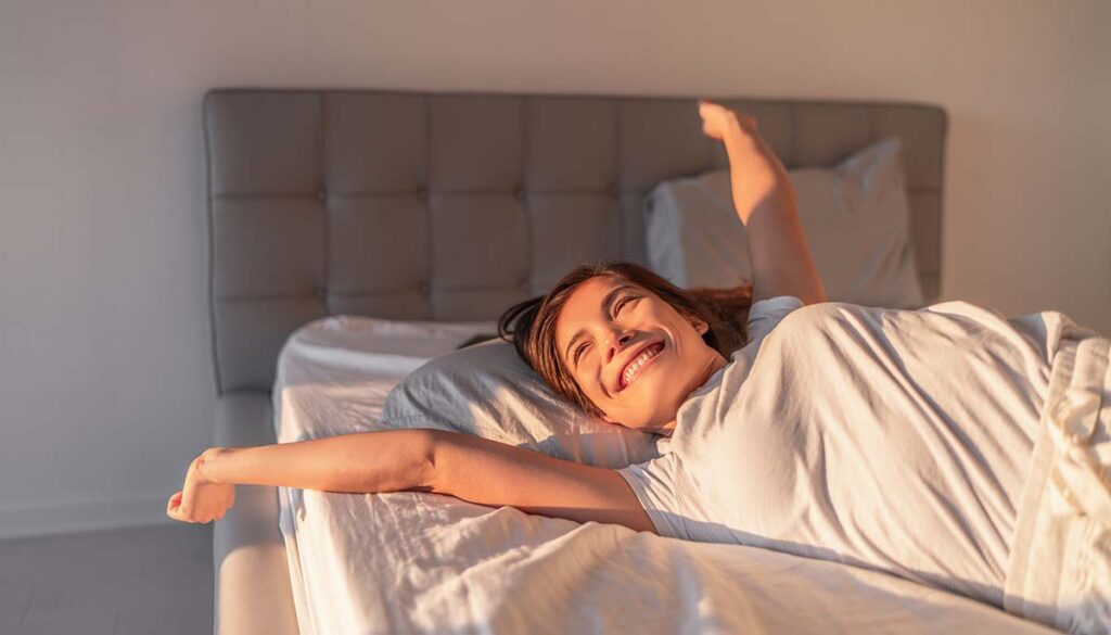 Happy girl waking up in the morning sunshine looking at sunrise sun in window excited to enjoy the day. Wake up energetic Asian woman lying in bed well rested from a good night sleep.