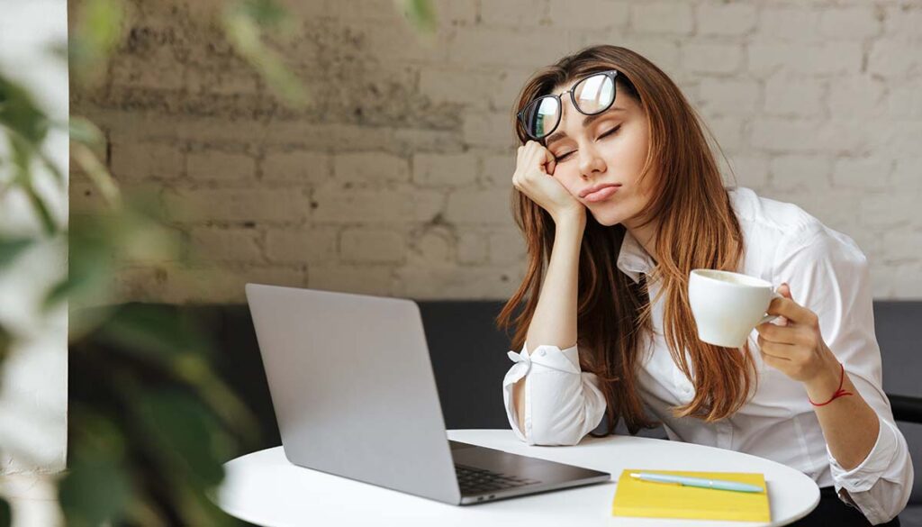 Portrait of a tired young businesswoman sitting at the table with laptop computer while holding cup of coffee and sleeping at a cafe