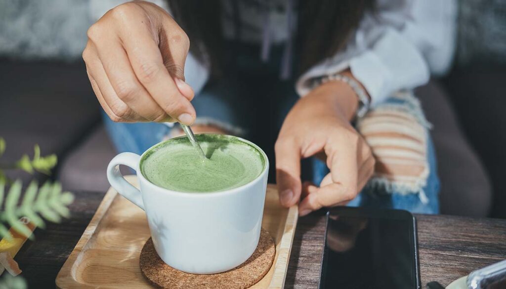 Woman holding spoon and stirring hot green tea matcha latte at cafe.