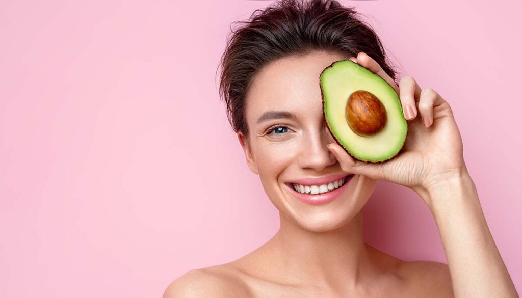 woman holds half an avocado in front of her face. 