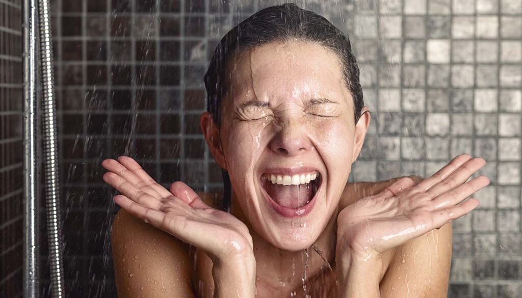 woman reacting in shock to cold shower water as she stands under the shower head washing her hair