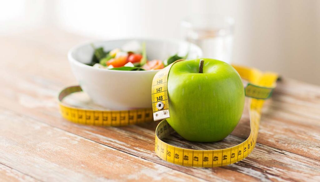 diet, healthy eating, food and weigh loss concept - close up of green apple and measuring tape with salad on wooden table