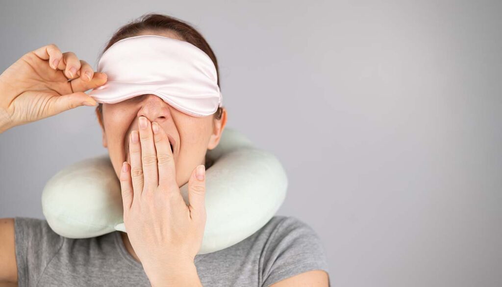 woman yawning in sleep mask and neck pillow against gray background