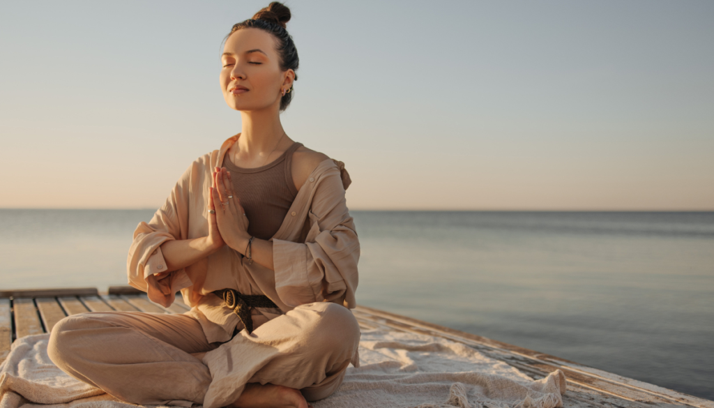 Woman sitting on a pier cross legged with eyes closed and hands pressed together