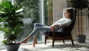 woman at home sitting on modern chair near window relaxing in living room with house plants