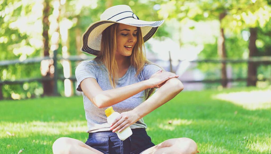 woman wearing a hat and putting on sunscreen in a park